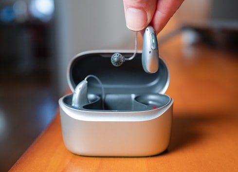 Rechargeable hearing aid held by fingers over its charging case.